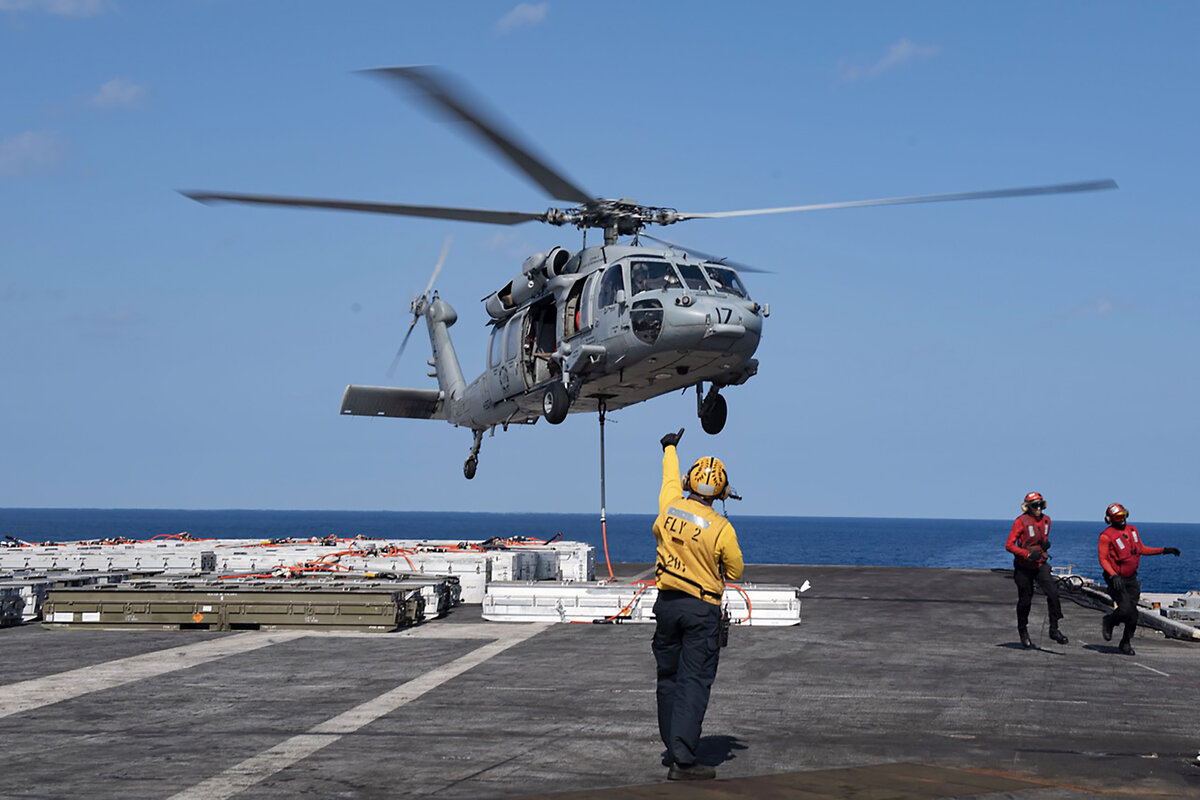 A crew member signals to a rising helicopter, from the deck of an aircraft carrier.