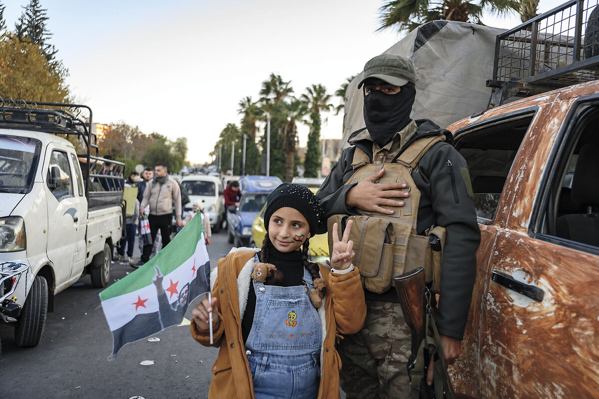 A young girl dressed in overalls and carrying a Syrian flag holds her fingers in a peace sign while posing for a photo.
