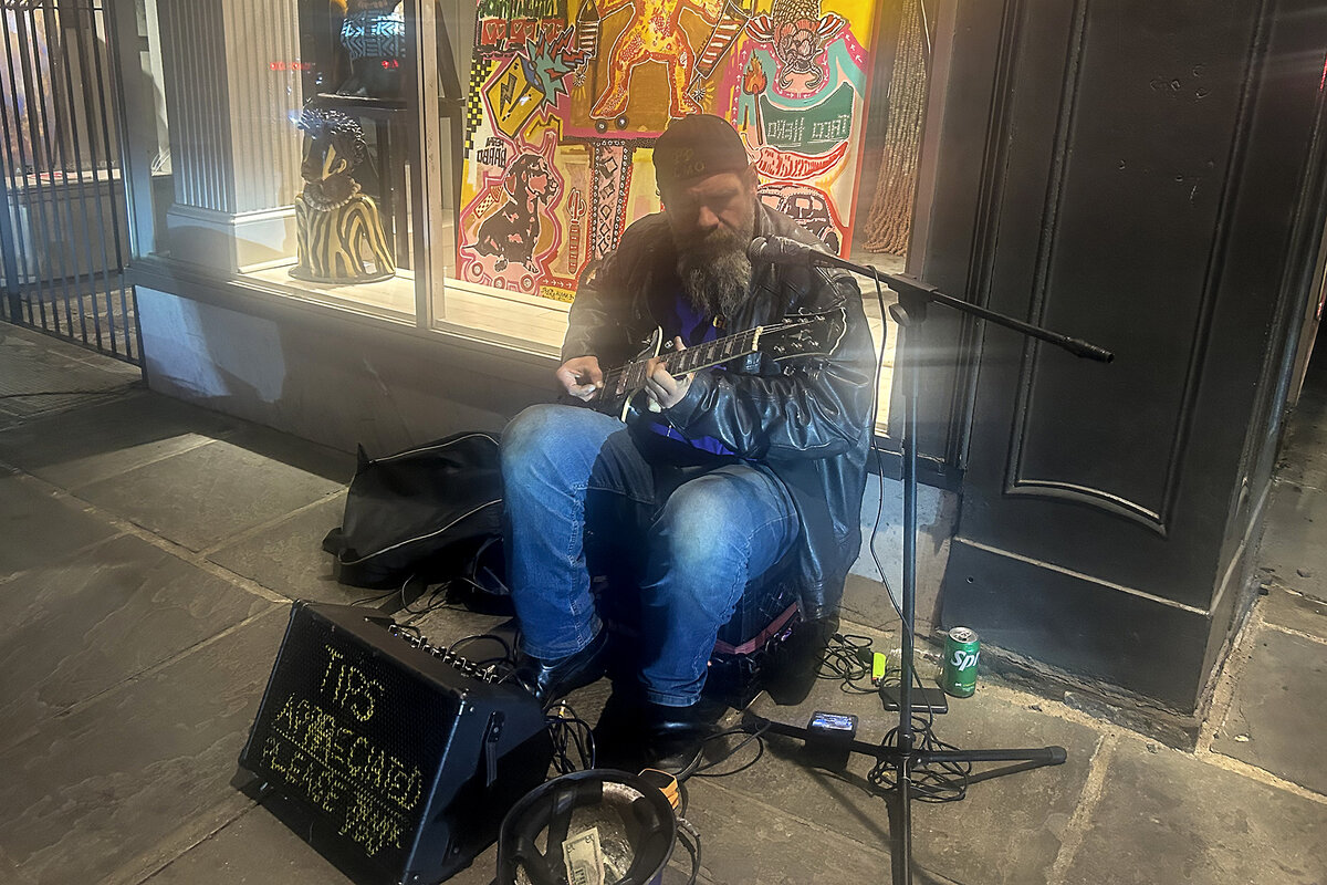 A man in jeans and a backward baseball cap sings and plays guitar in New Orleans' French Quarter. His speaker says "Tips Appreciated."