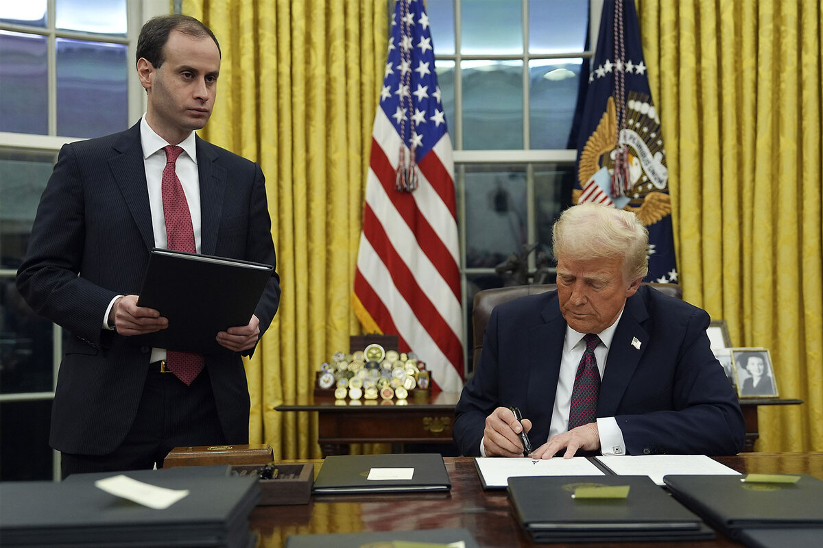Donald Trump sits at his desk in the White House Oval Office, with an American flag in the background, and signs an executive order.