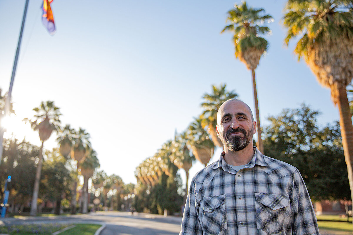 Judd Ruggill, head of the department of public and applied humanities at the University of Arizona, stands facing the camera and smiles on a street bordered by palm trees.