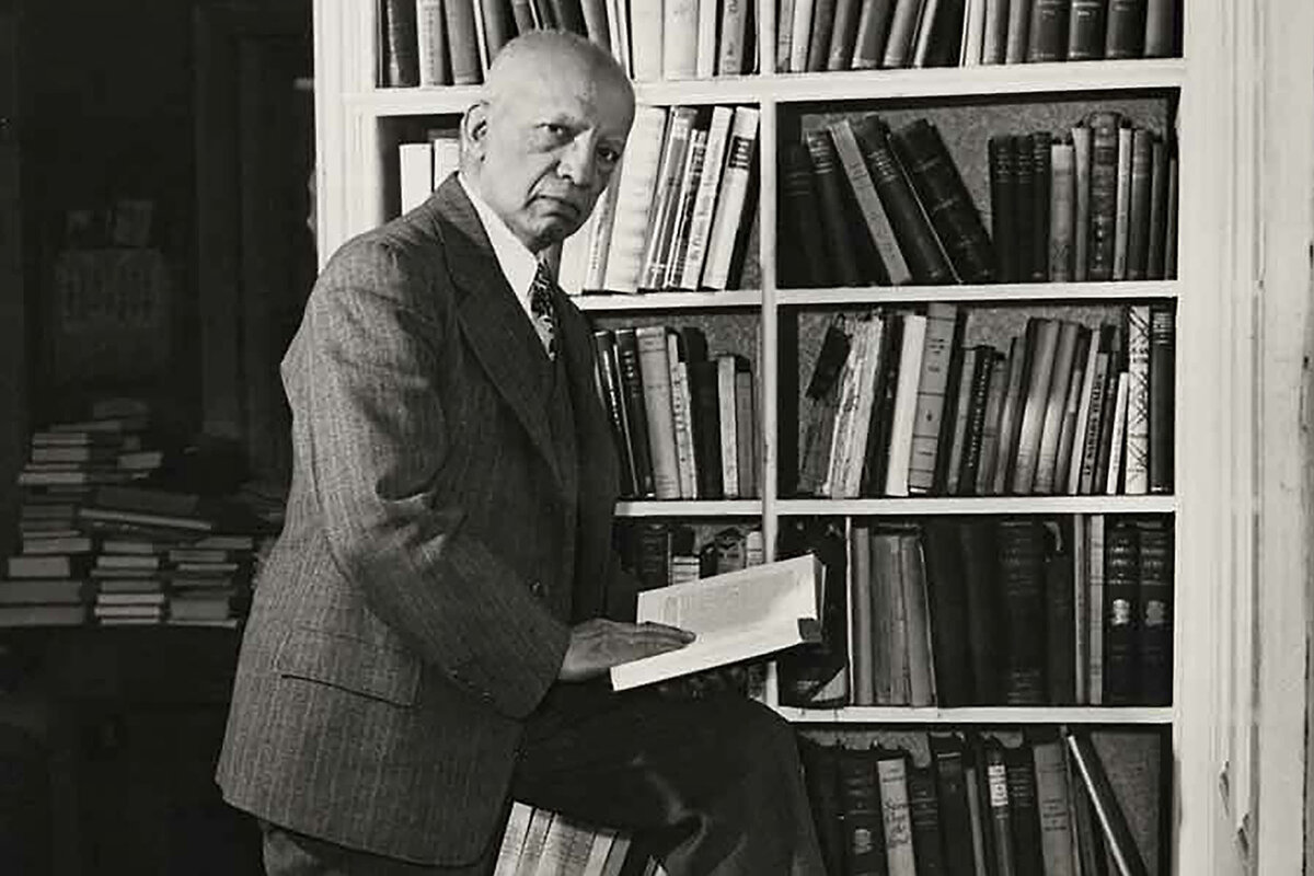 In a black-and-white photo, historian and author Carter G. Woodson stands next to a bookshelf in a suit looking at the camera while holding a book open.