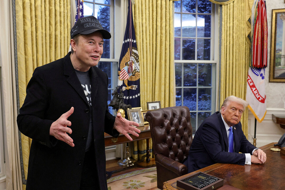 Elon Musk, wearing a baseball cap, gestures as he speaks next to U.S. President Donald Trump, who sits at his desk in the Oval Office of the White House.