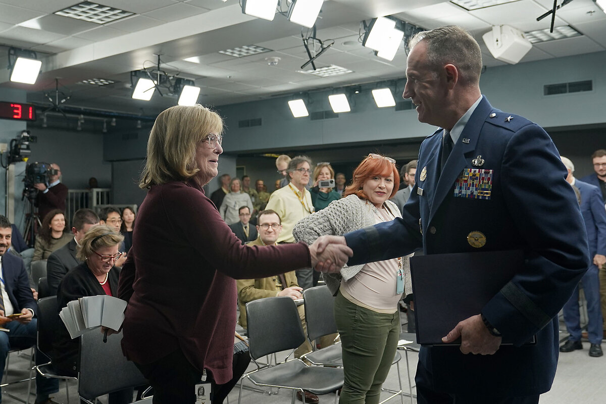Other reporters look on in the briefing room as Pat Ryder, in uniform, shakes hands with journalist Barbara Starr.