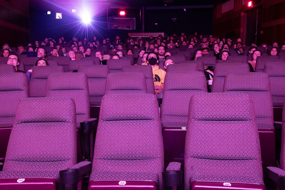 Moviegoers partially fill a theater. The light of the projector can be seen over their heads.