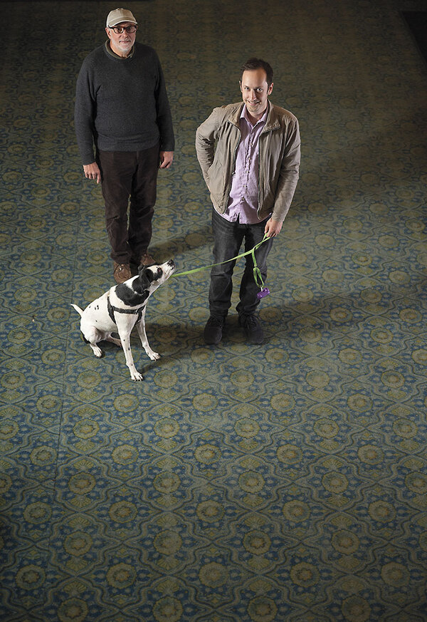 Two men stand on an intricate theater lobby carpet with a dog on a leash.