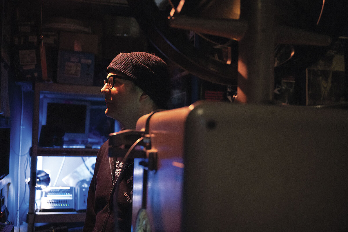 A projectionist keeps an eye on a movie from a darkened projection booth.