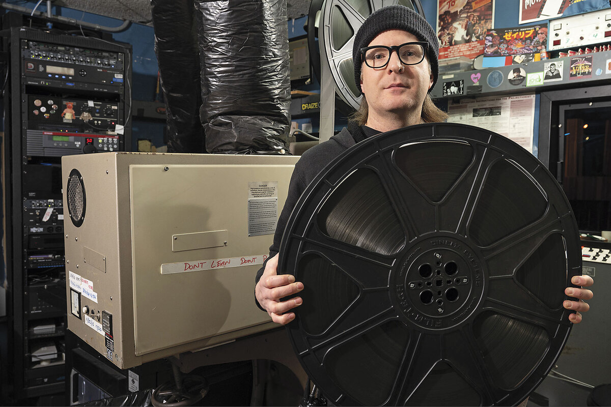 A man wearing glasses and a winter cap poses in a crowded projection booth.