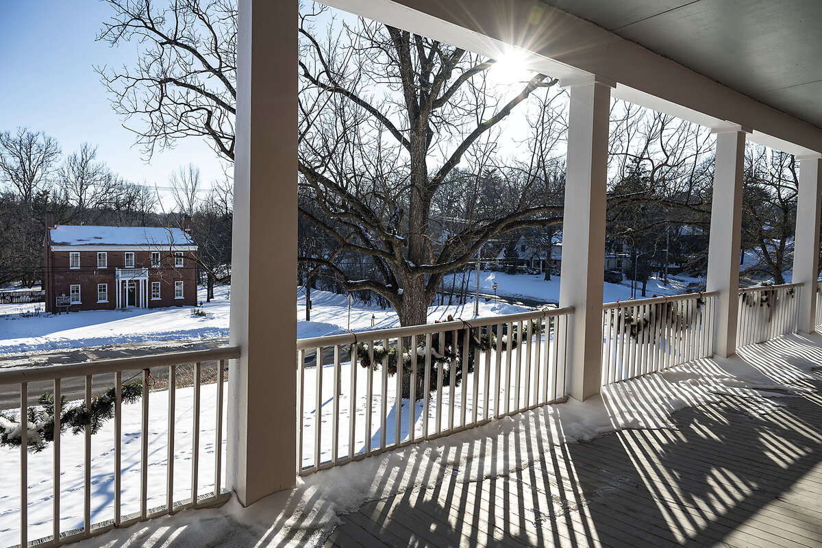 A brick building stands in the distance as sunlight streams onto a pillared porch.