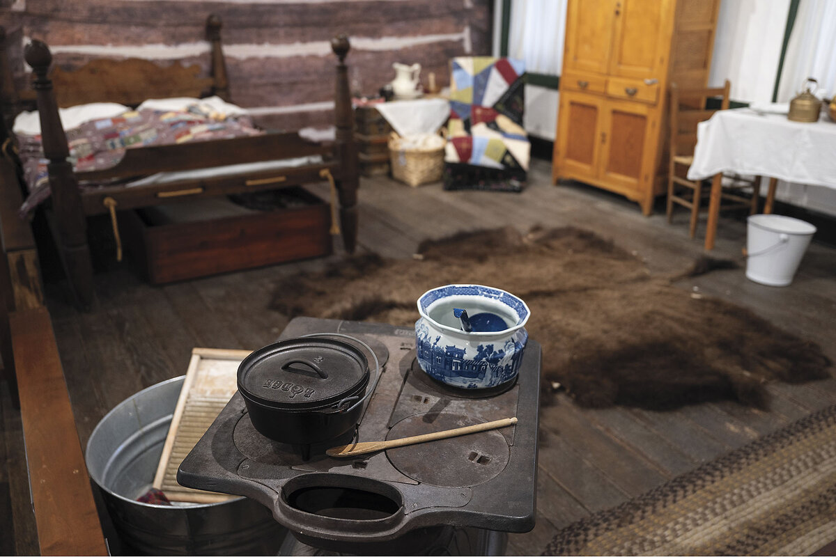 Pioneer-era cooking utensils sit on a washboard and basin in front of a four-poster bed and wardrobe.