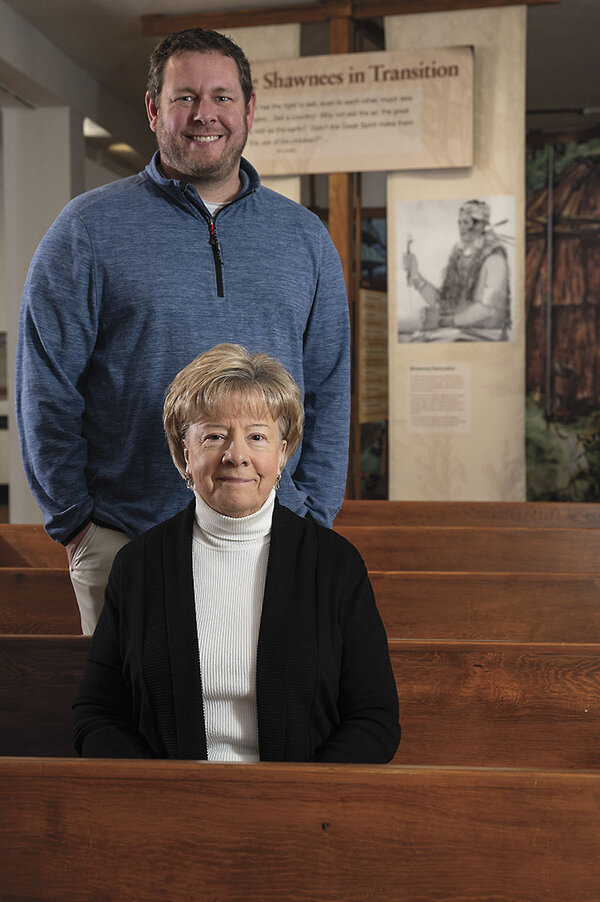 A woman sits in a chapel pew with a man standing behind her.