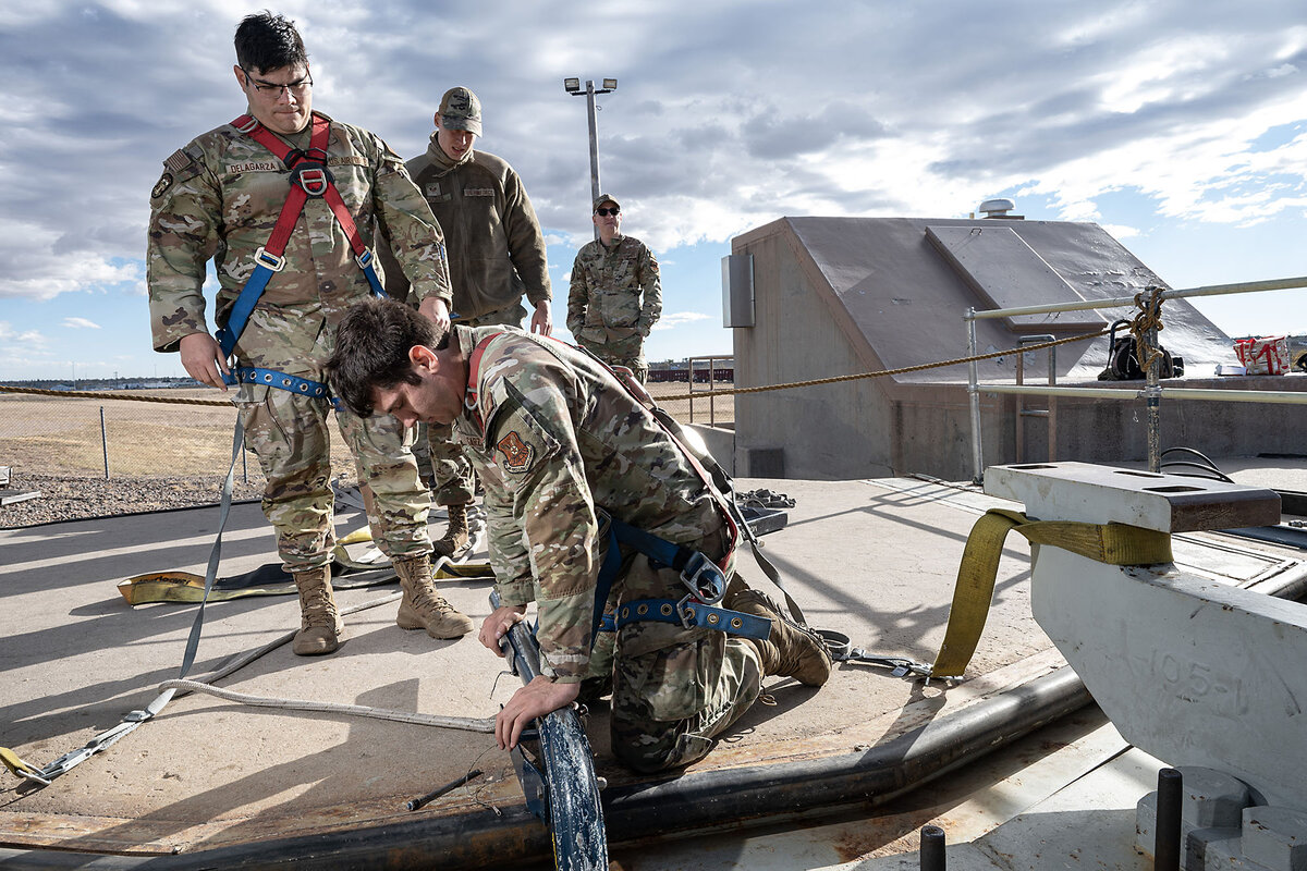 A maintainer kneels as he leans on a metal bar. Three others watch on.