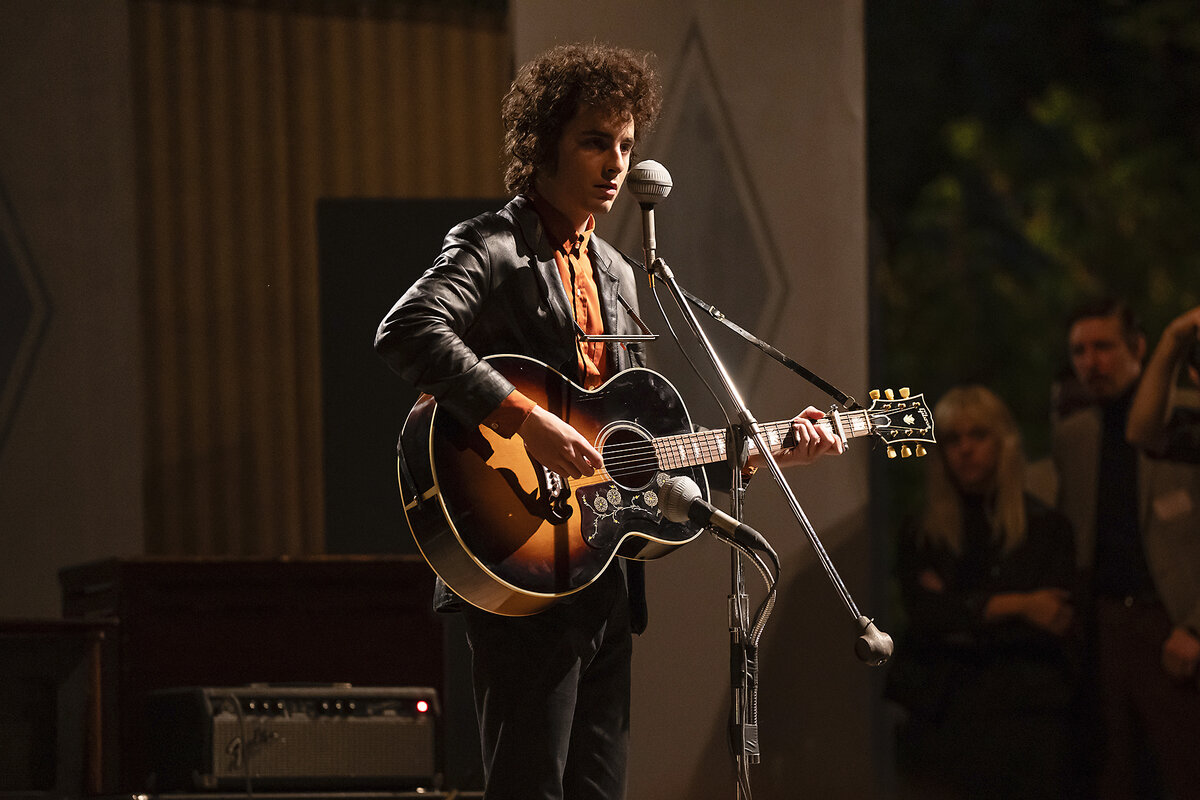 Actor Timothée Chalamet, portraying Bob Dylan, stands at a microphone holding a guitar