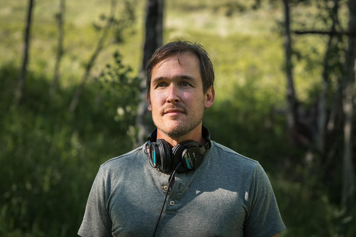 Journalist Nick Mott, with headphones wrapped around his neck, is pictured in front of small trees in southwest Montana in the summer of 2024.
