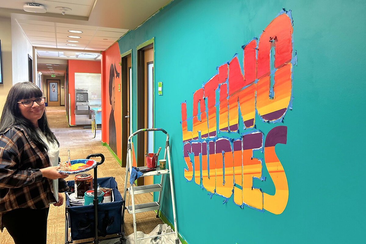 Artist Cindy Hernandez stands in front of a mural holding paints and looking at the camera smiling in Austin, Texas