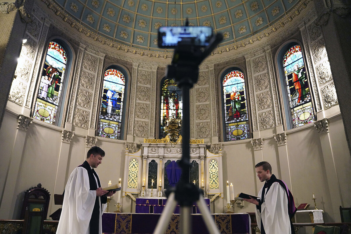 Two clergy members stand in front a tripod capturing video for a remote congregation, with stained-glass windows behind them.