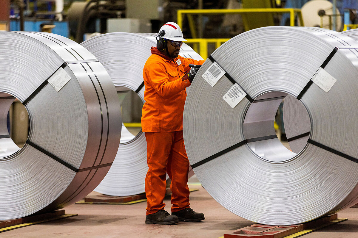An orange-suited worker stands next to large bundles of flat-rolled steel.