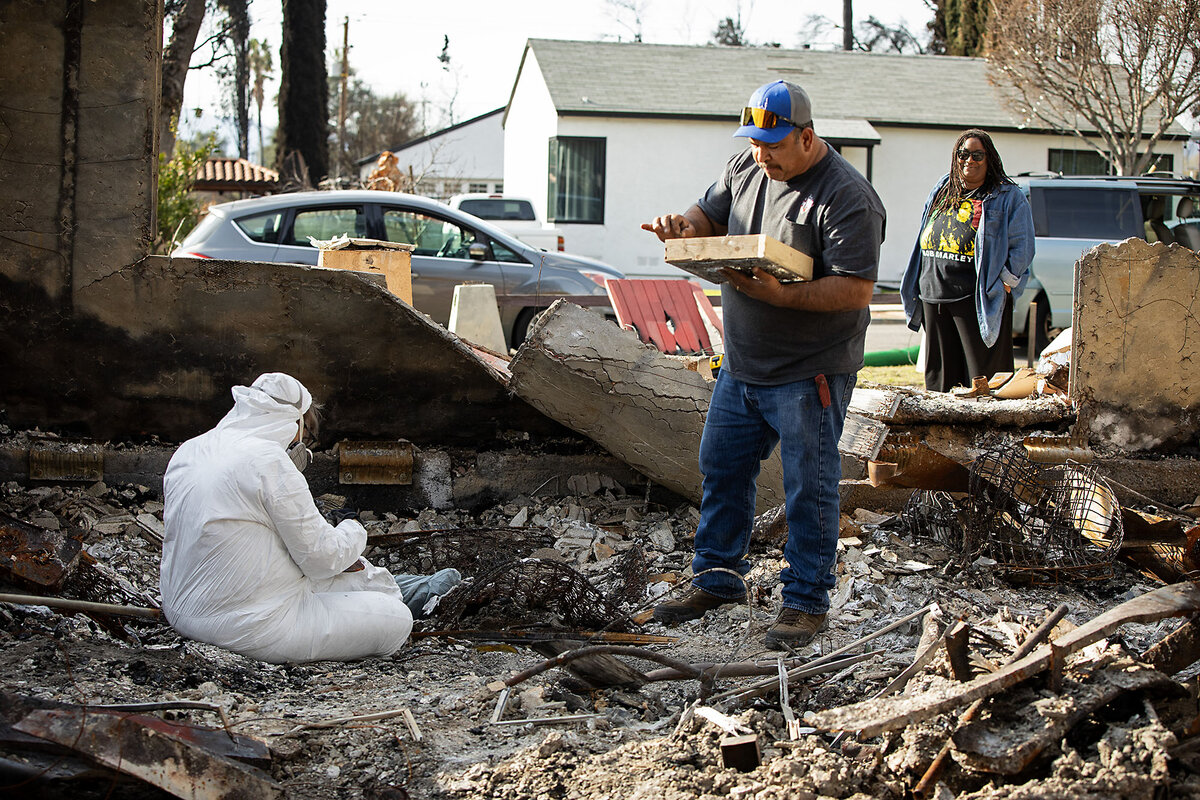Nicole Moore, wearing a white hazmat suit, sits in the ashes sifting for belongings while neighbor Jose Castillo stands and helps.