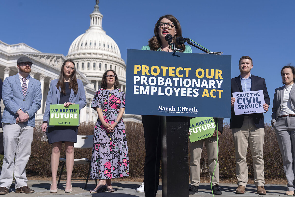 Standing in front of supporters with signs, Rep. Sarah Elfreth speaks to reporters. The Capitol dome is seen behind them. 