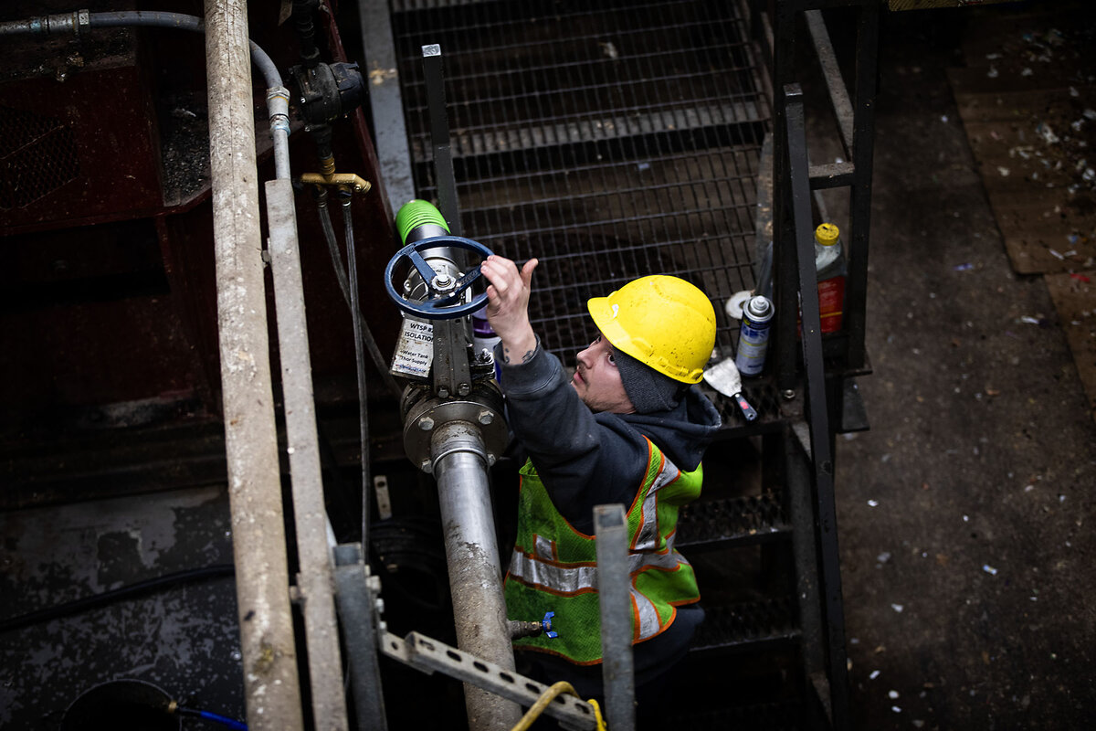 A man in a yellow hard hat and safety vest turns a valve attached to a series of pipes that control water flow.