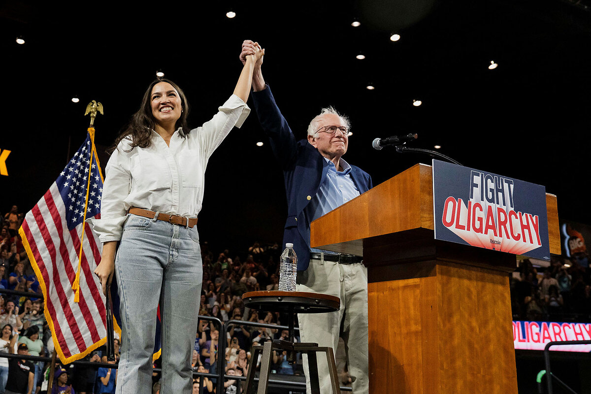 Rep. Alexandria Ocasio-Cortez and Sen. Bernie Sanders join hands and smile in front of their large audience.