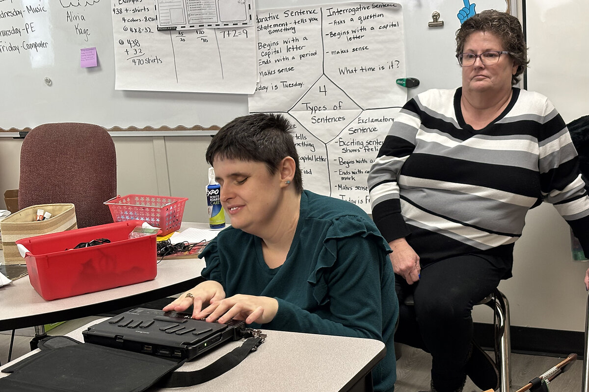 Delilah Willis sits at a desk and reads students a book using braille while her mother, Deanna Willis, watches
