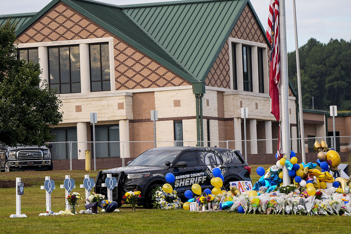 An outdoor memorial near a school flagpole includes crosses, flowers, and blue and yellow balloons.