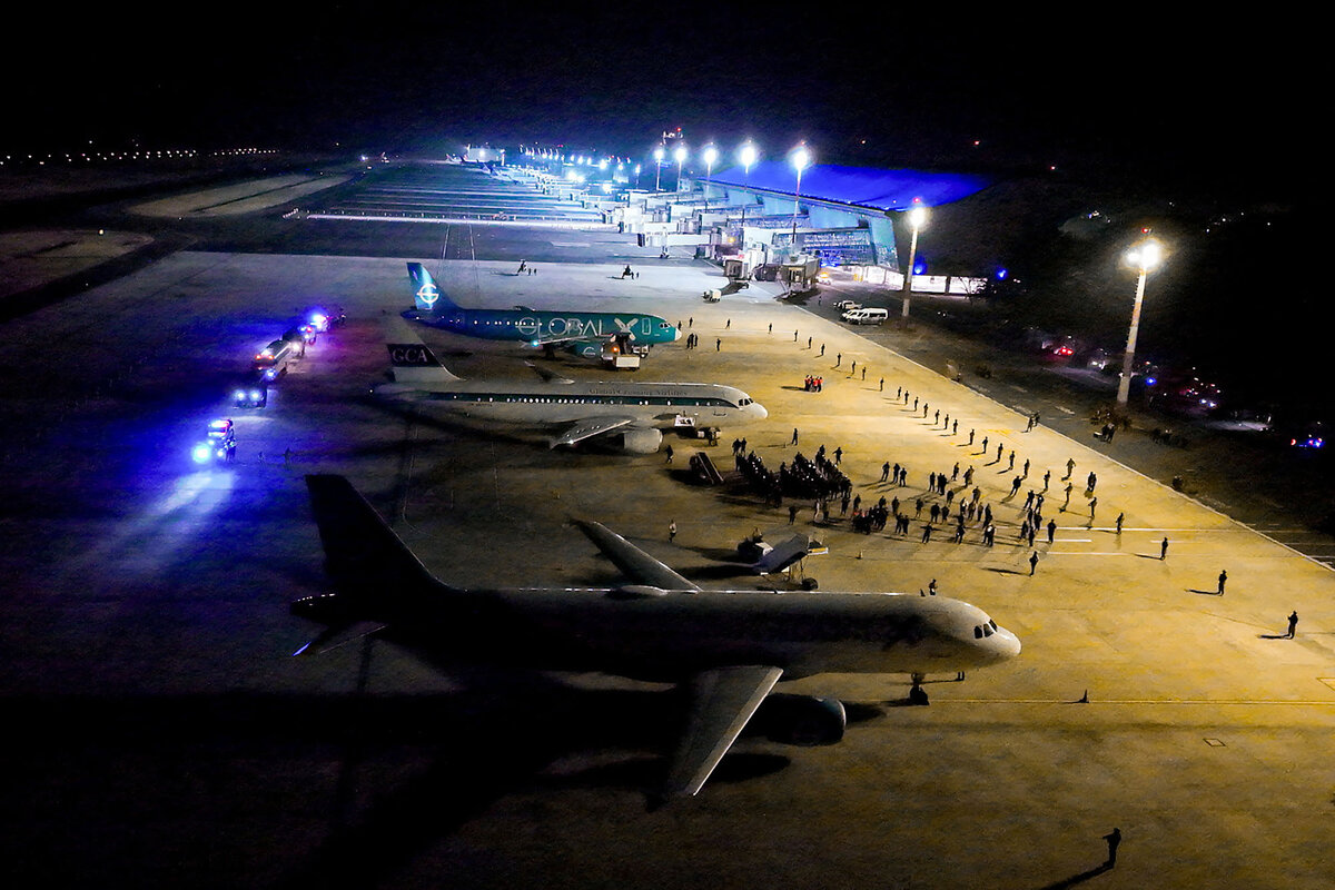 An overhead view shows people gathering near three airplanes, at night at the El Salvador International Airport.