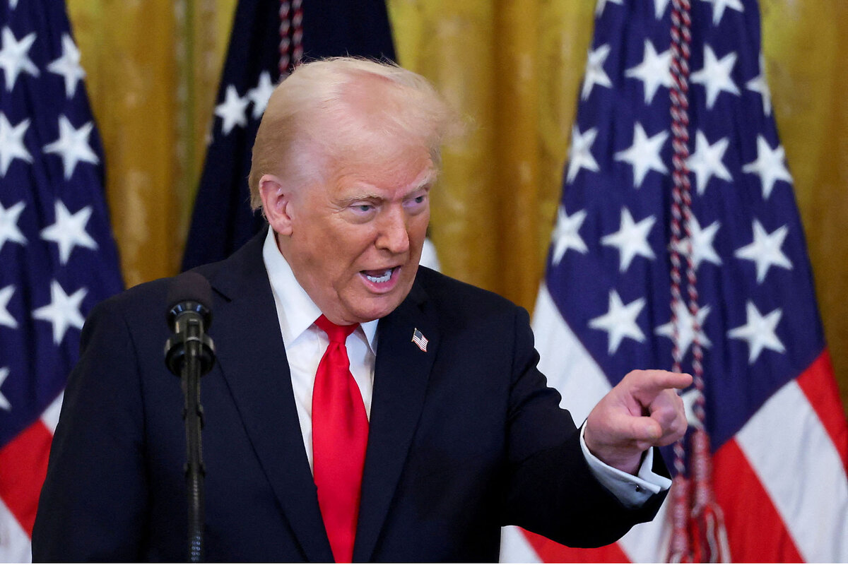 Standing in front of three American flags, President Donald Trump points at someone while he is talking. 