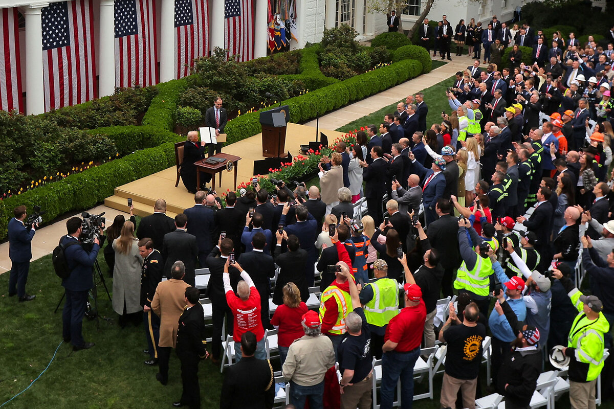 Reporters and others watch as President Trump, seated at an on-stage desk, holds his executive order on tariffs.