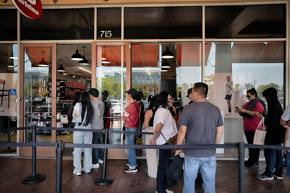 A line of shoppers stand outside the glass doors of New Balance shoe store, waiting for their turn to go in.