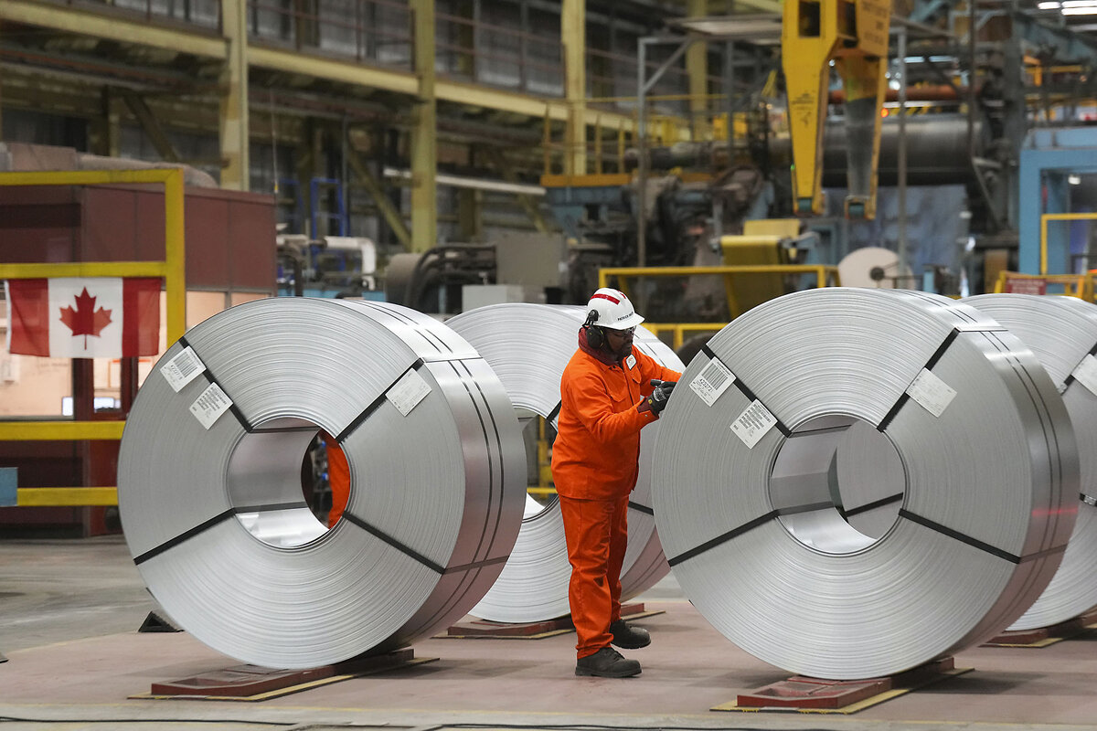 An orange-suited steel worker stands on a shop floor next to coils of flat-rolled steel.