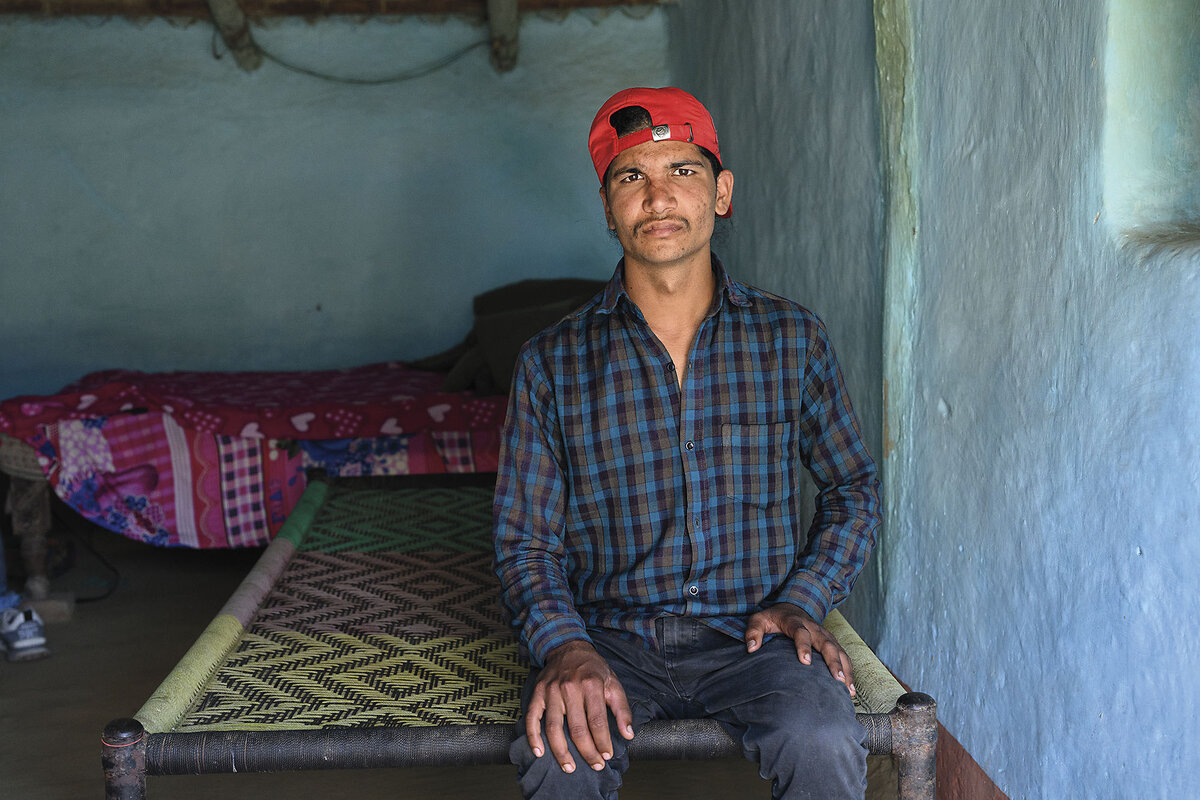 A man in a plaid shirt and red baseball cap sits in a sparse bedroom.