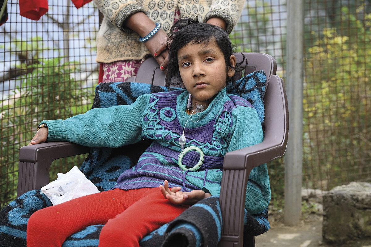 A young girl sits on a chair with a woman standing behind her.