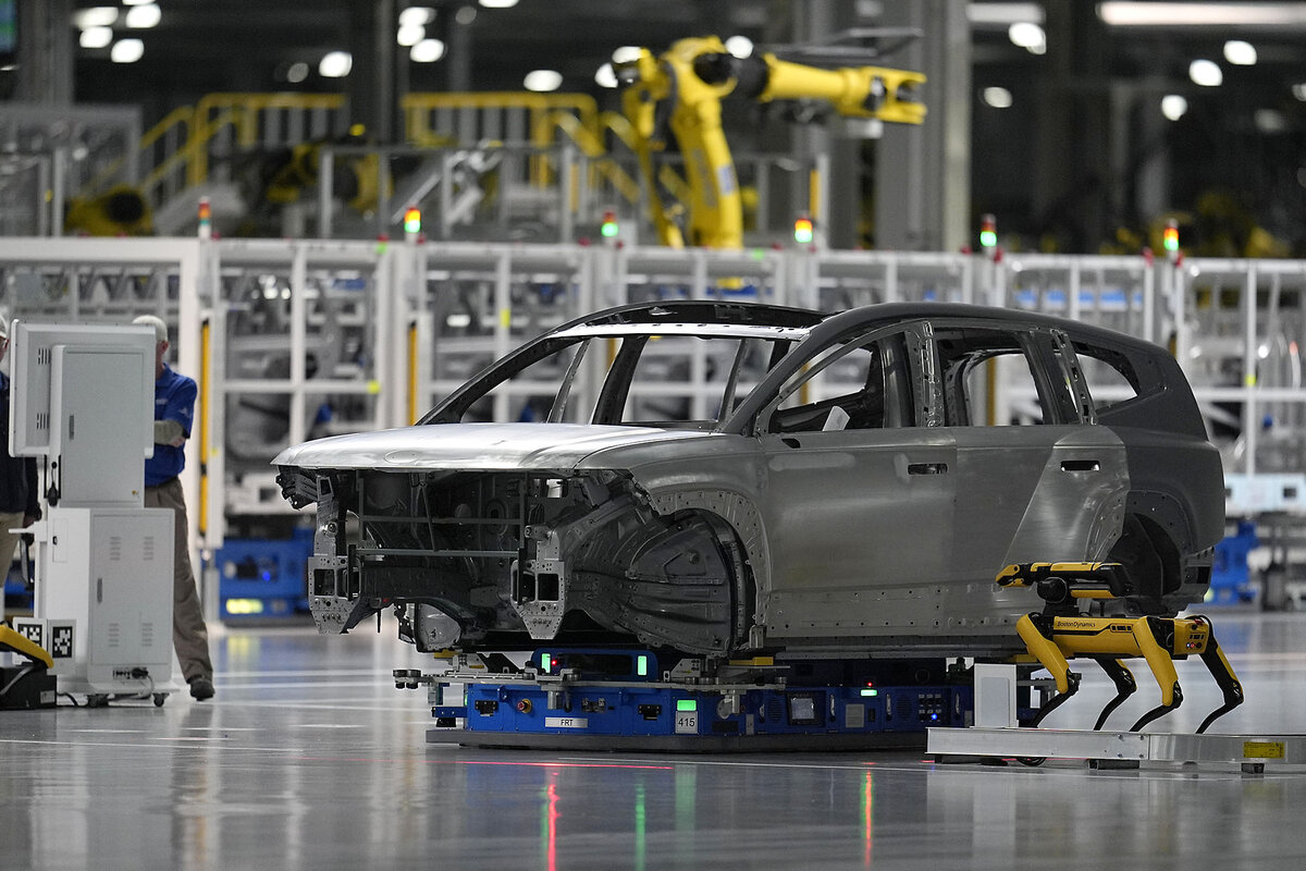 The shell of a gray car, hoisted up on lift, has open doors out of which robotic yellow arms, extended for action, can be seen.