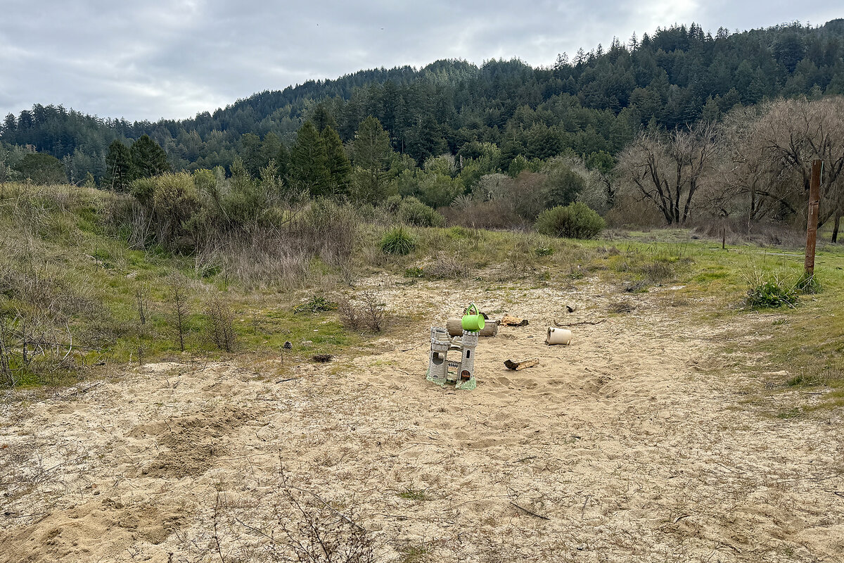 Play castles and plastic toys lie in a former sand trap of the rewilded San Geronimo Commons. The former golf course is now a nature preserve. Behind the sand are trees and hills.