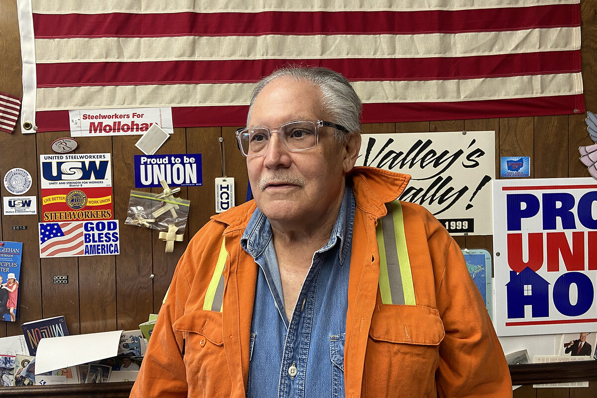 Mark Glyptis wears an orange steelworker's jacket, with pro-labor signs and a U.S. flag behind him at the local union's office.