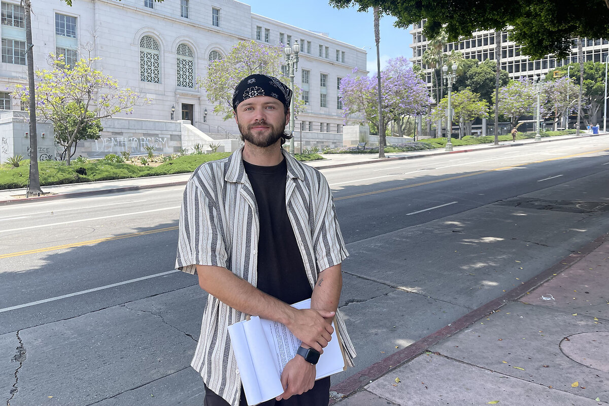Paul Nauta, wearing a black T-shirt and carrying a small white sign, stands in front of a white building.