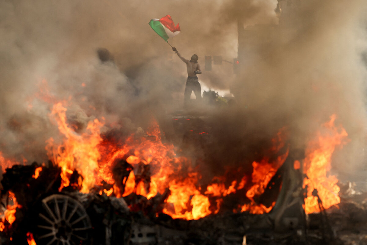 A man waves a Mexican flag as smoke and flames rise from a nearby burning vehicle.