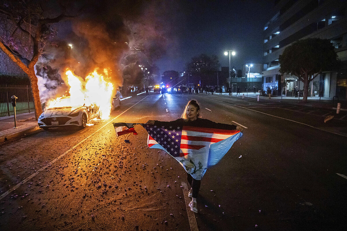 A person carrying a flag like a cape walks on a dark street past a burning car during protests over immigration raids in Los Angeles