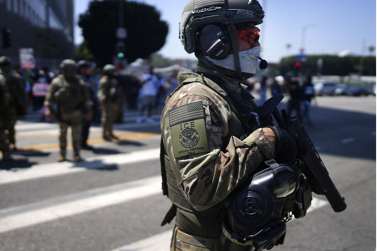 An armed and helmeted ICE Special Response Team member stands guard while protesters gather outside