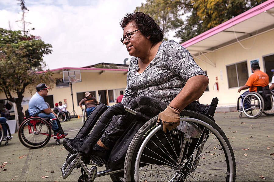 Woman in wheelchair in outdoor courtyard, with other wheelchair users in background.