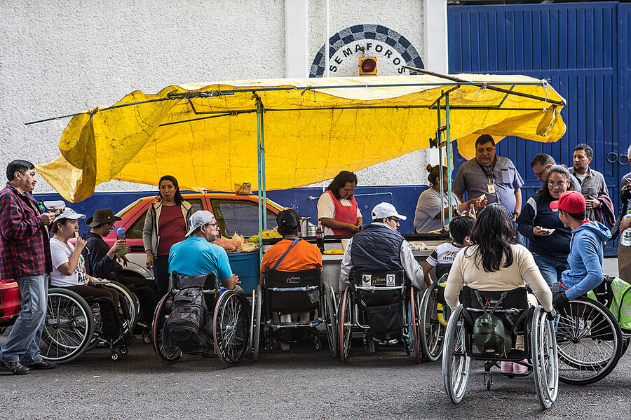 Group of wheelchair users gather under a yellow tarp outside while eating.