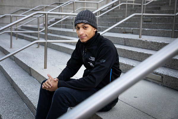 William Brown, slightly smiling and wearing a long-sleeve shirt, dark-colored pants and a knit ski cap, sits outdoors on a set of wide stairs in Boston. Mr. Brown is a shop steward for Unite Here Local 26 and helped lead a strike in 2024 that resulted in a new contract for workers at the Omni hotel where he works. 