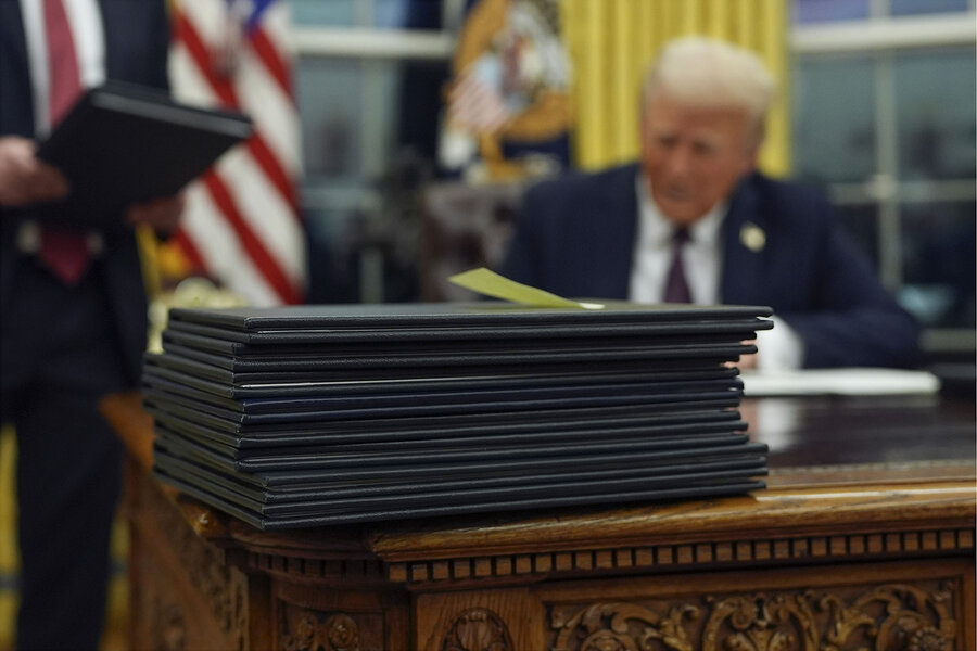 A stack of executive orders in black folders sit on a desk, with President Trump behind them signing another one.