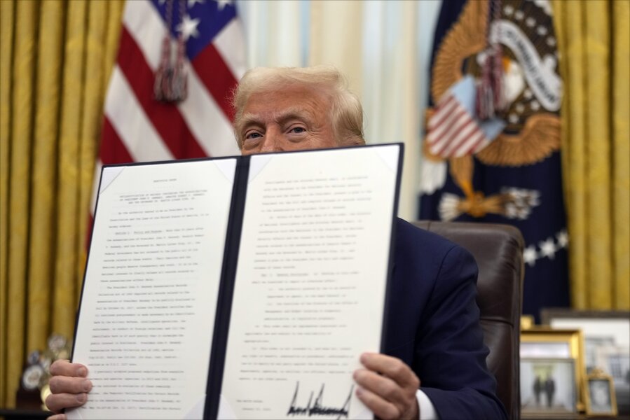 President Donald Trump holds an executive order in the Oval Office of the White House. The document blocks the lower half of his face.