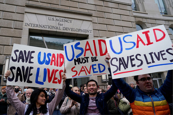 Protesters stand and hold signs outside the USAID building, including one sign that reads USAID saves lives.