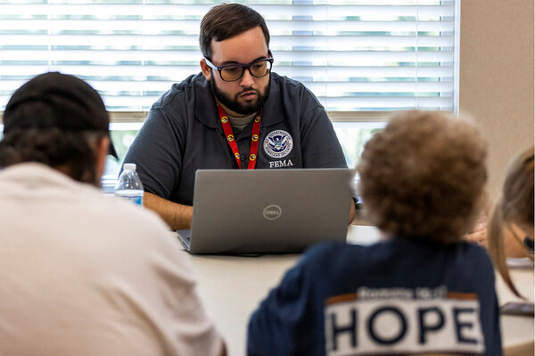 A man wearing a blue FEMA shirt scans his computer for information to give two people, sitting across from him.