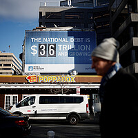 As a pedestrian walks by, a sign is visible on a building, with $36 trillion listed as the national debt.