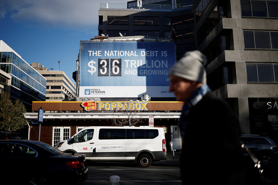 As a pedestrian walks by, a sign is visible on a building, with $36 trillion listed as the national debt.